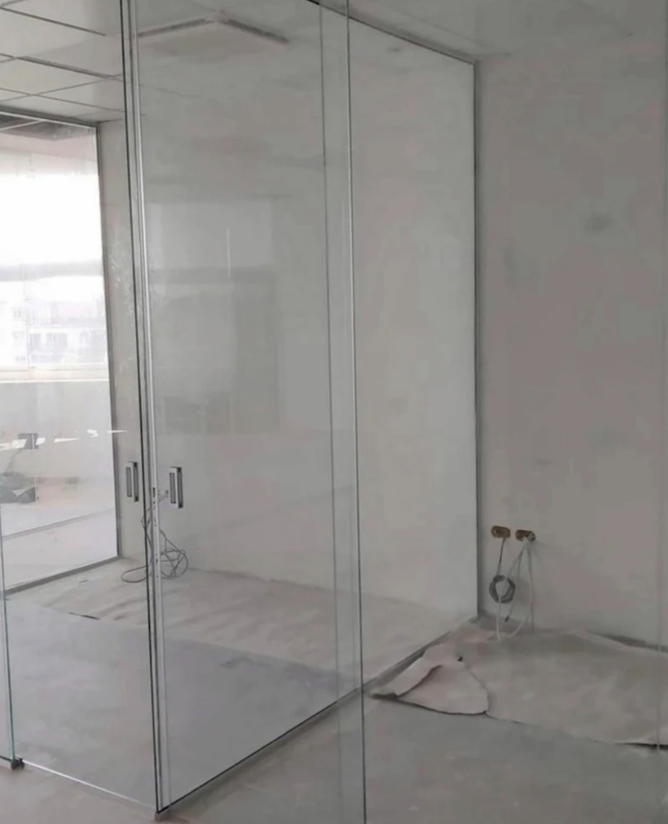 Nowoczesne, szklane drzwi przesuwne z metalowymi uchwytami, oddzielające pomieszczenia w surowym wnętrzu. Widoczne przyłącza instalacji na ścianie.