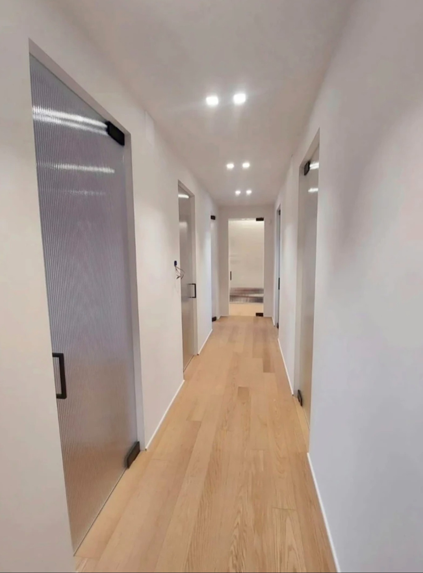 Nowoczesny korytarz z drewnianą podłogą i szklanymi drzwiami z czarnymi uchwytami. Minimalistyczny design, jasne oświetlenie.