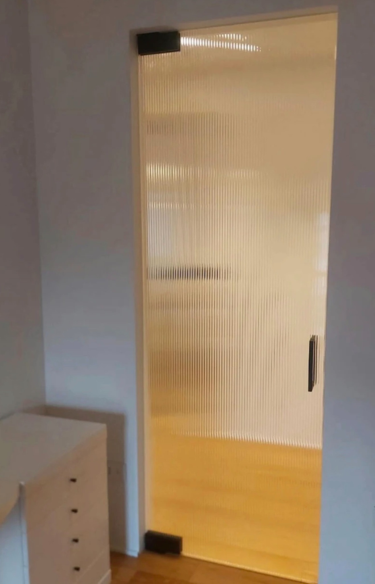 Matowe drzwi z ryflowanego szkła z minimalistyczną klamką, zamontowane w jasnym wnętrzu. Widoczny fragment komody i drewniana podłoga.