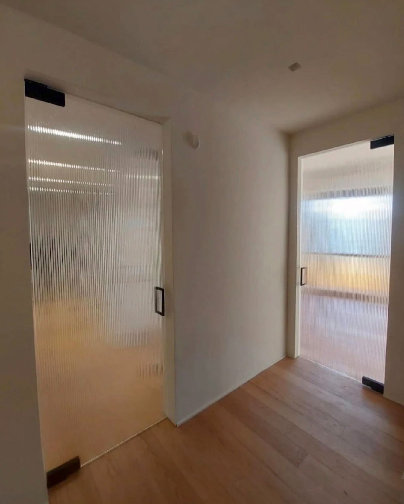 Dwa wejścia z prążkowanymi drzwiami szklanymi, czarne okucia, jasne drewniane podłogi, minimalistyczny design, neutralna kolorystyka ścian.