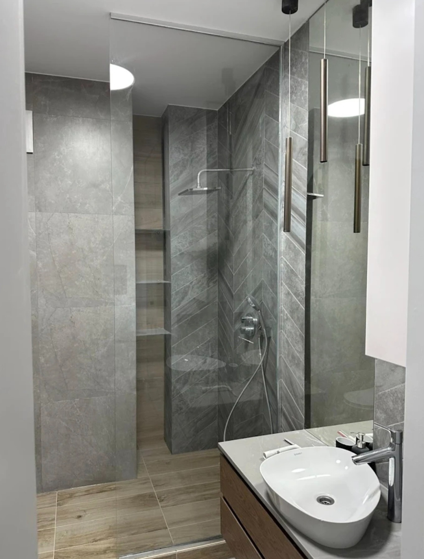 Nowoczesna kabina prysznicowa ze szklaną ścianą, szare płytki w jodełkę, umywalka nablatowa i minimalistyczne oświetlenie w łazience.