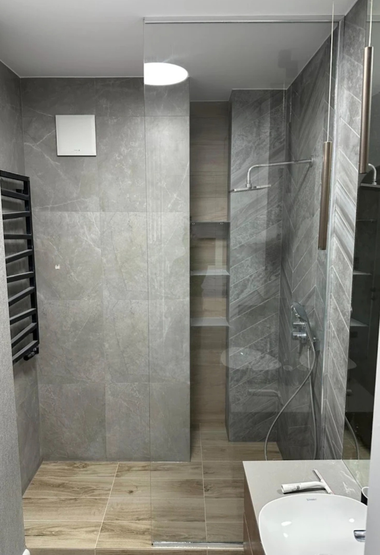 Nowoczesna kabina prysznicowa z pojedynczą taflą szkła, szare płytki imitujące marmur, grzejnik drabinkowy i drewnopodobna podłoga. Minimalistyczny design łazienki.