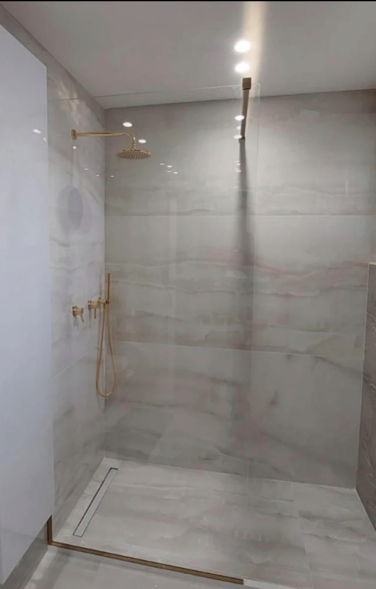 Nowoczesna kabina prysznicowa z elementami w kolorze złotym, wykończona płytkami imitującymi marmur. Widoczna szklana ścianka i deszczownica.