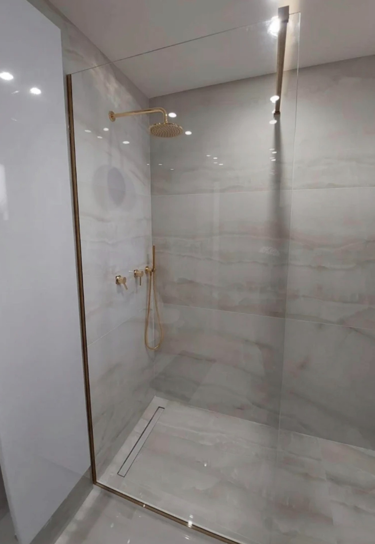 Nowoczesna kabina prysznicowa ze złotymi elementami armatury i szklaną ścianką, widok z boku, minimalistyczny design, jasne płytki imitujące marmur.