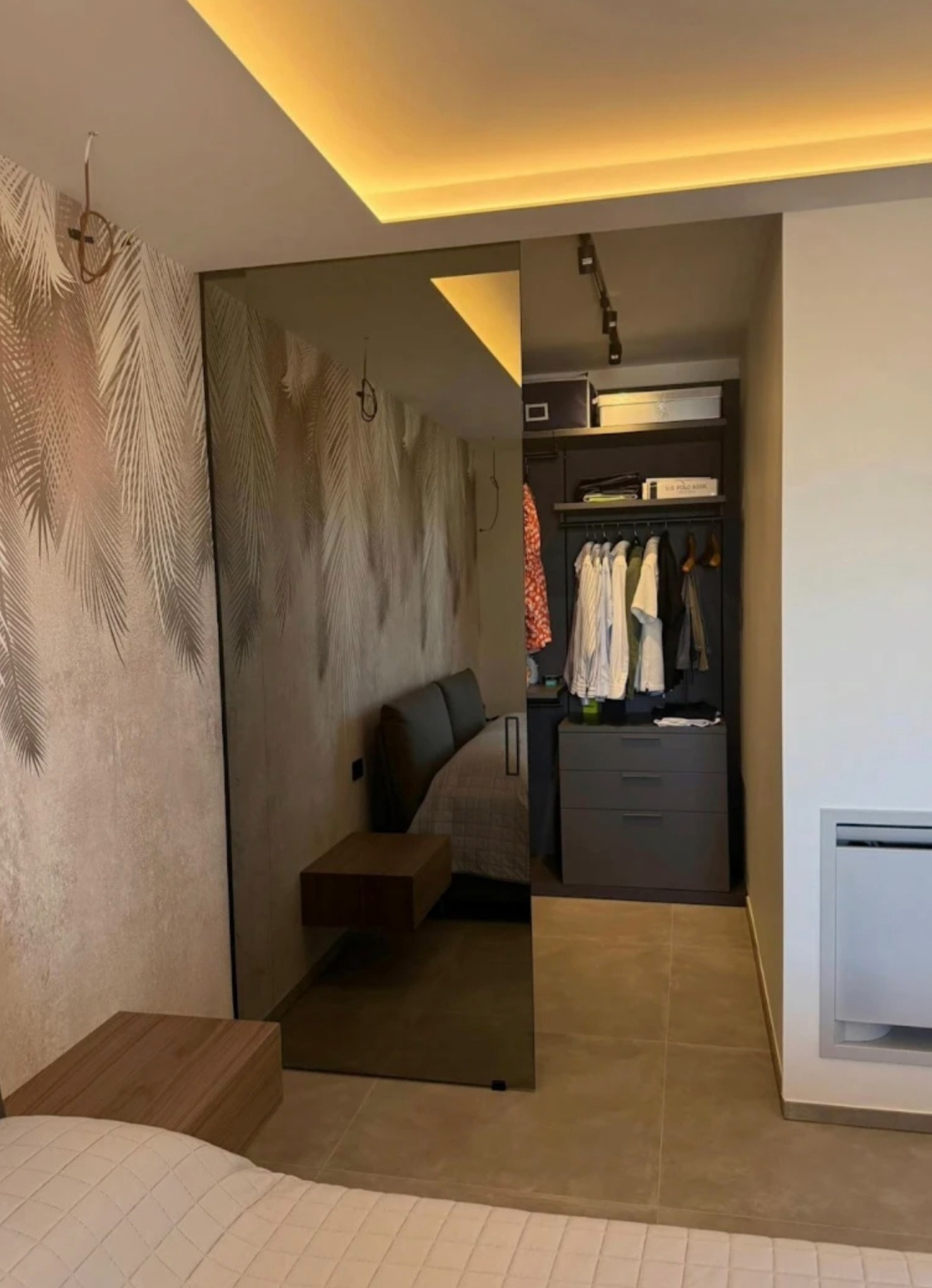 Szklane drzwi przesuwne oddzielają sypialnię od garderoby. Widoczne oświetlenie sufitowe i tapeta w liście. Minimalistyczny design wnętrza.