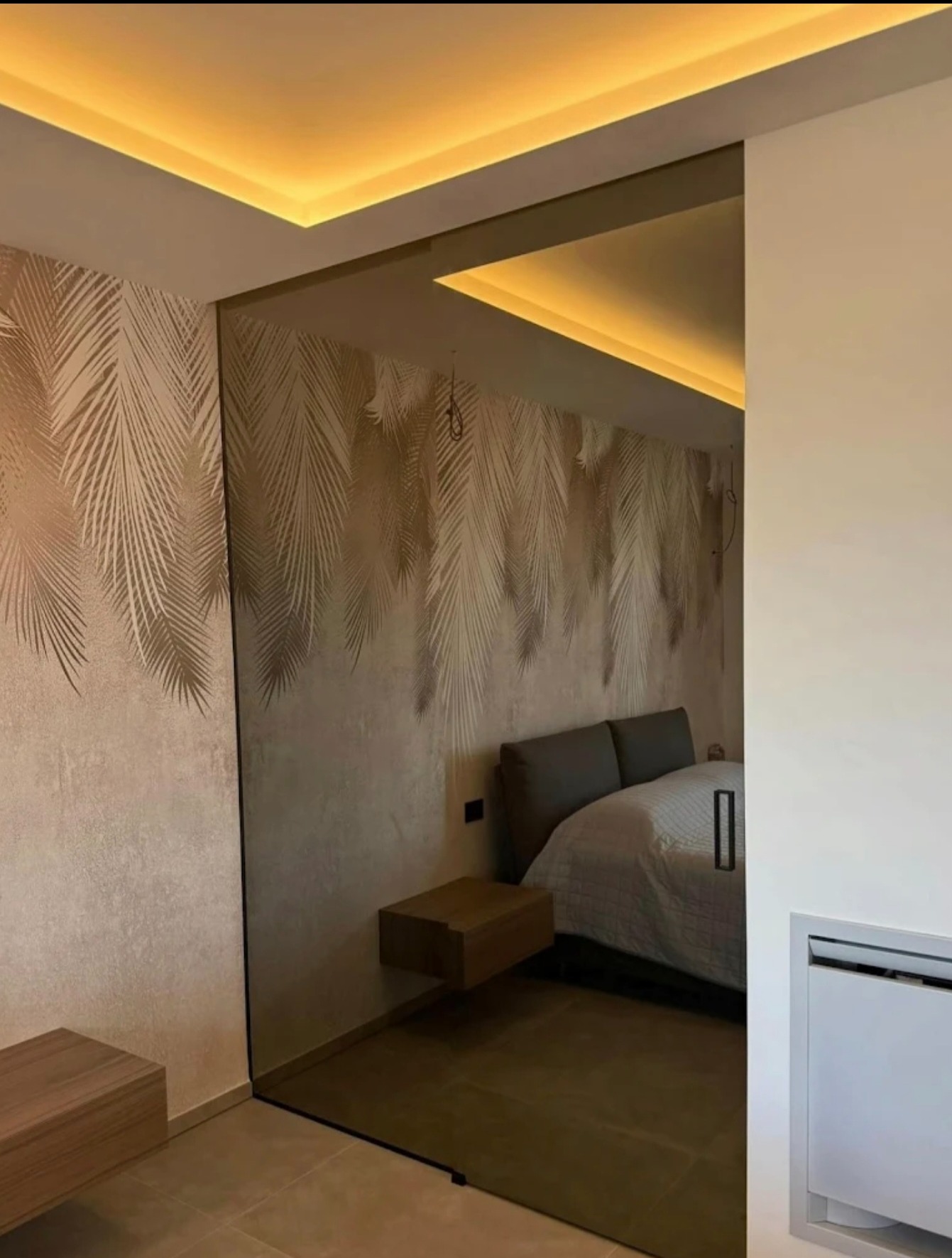 Nowoczesne drzwi przesuwne ze szkła w sypialni z tapetą w liście palmowe i podświetlanym sufitem. Minimalistyczny design, ciepłe oświetlenie.
