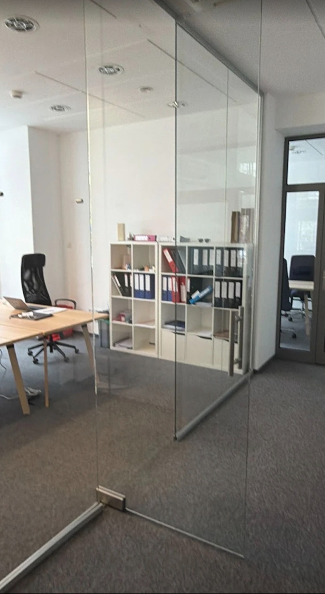 Szklana ścianka działowa w biurze z widocznymi okuciami i elementami mocującymi. W tle biurko, krzesło i regał z dokumentami.