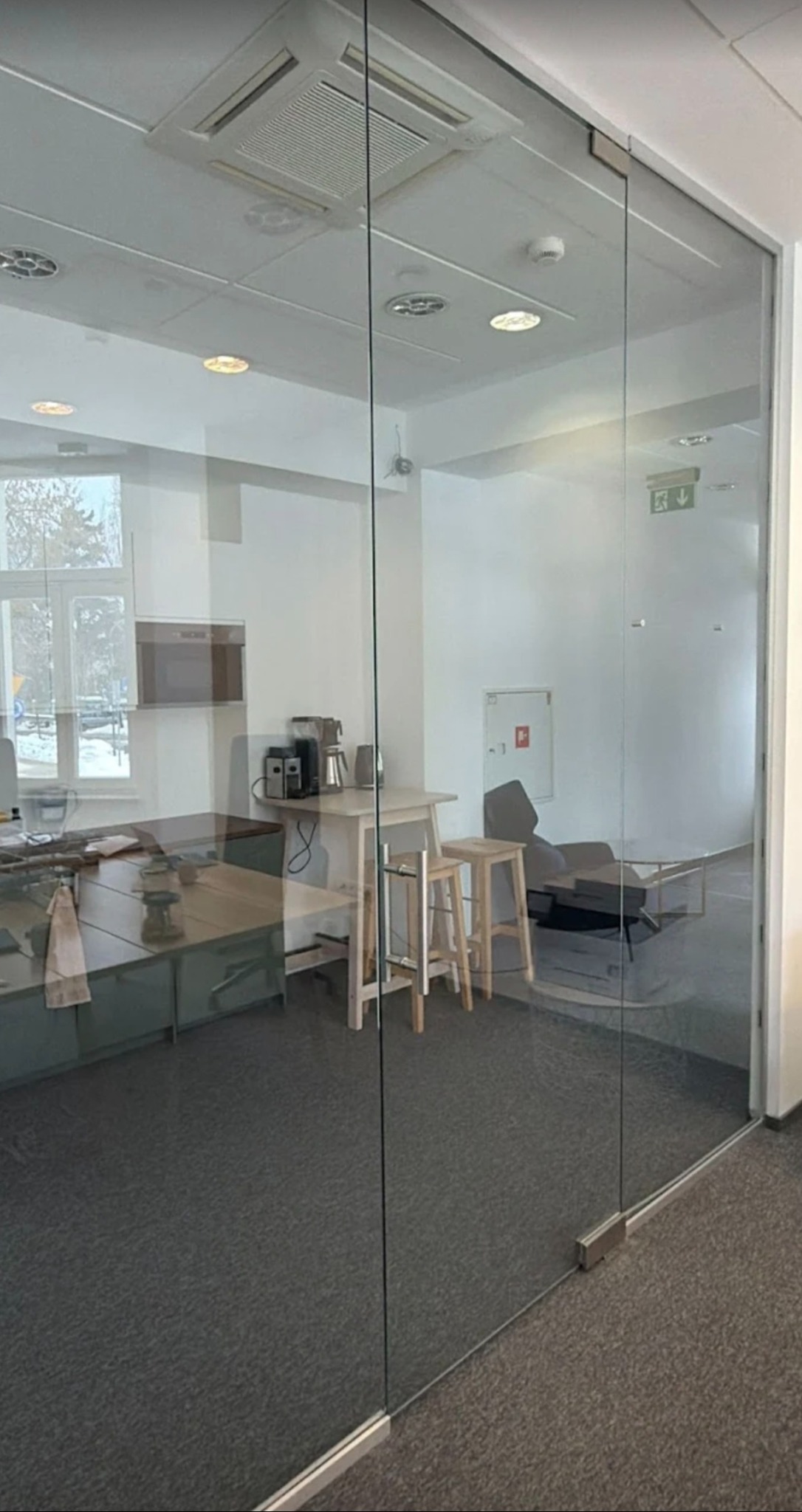 Szklane drzwi biurowe z widocznym wnętrzem: biurko, krzesła barowe, ekspres do kawy. Minimalistyczny design, jasne kolory, nowoczesne oświetlenie sufitowe.