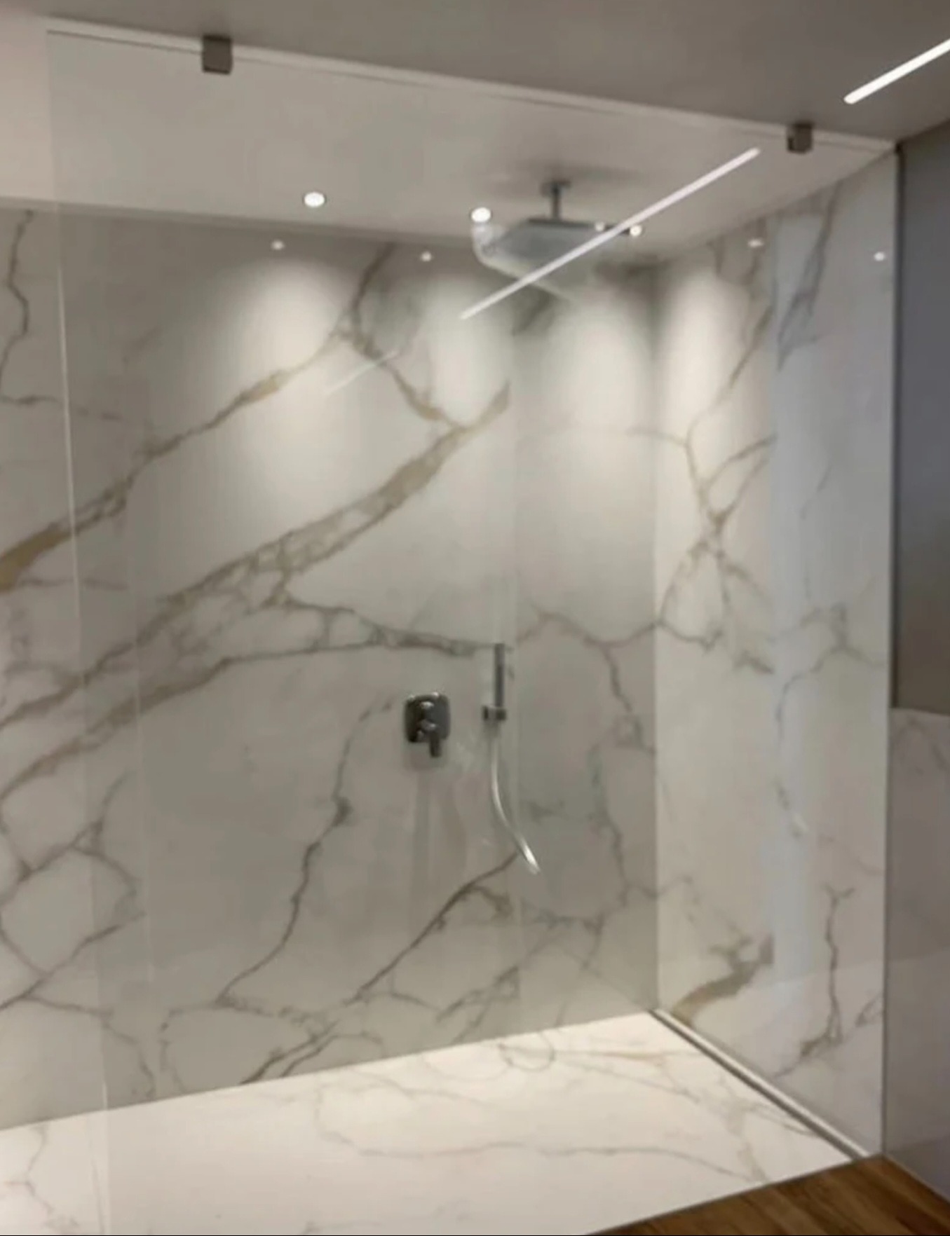 Nowoczesna kabina prysznicowa z marmurowymi płytami i szklaną ścianą, oświetlona punktowymi lampami sufitowymi. Minimalistyczny design, widoczna armatura.