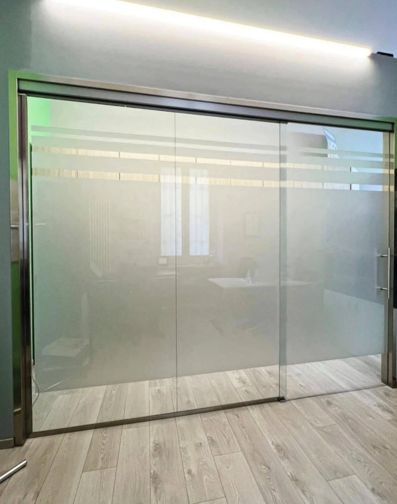 Przesuwne drzwi z matowego szkła z poziomymi paskami, osadzone w metalowej ramie, na tle jasnego wnętrza biura z drewnianą podłogą i oświetleniem LED.