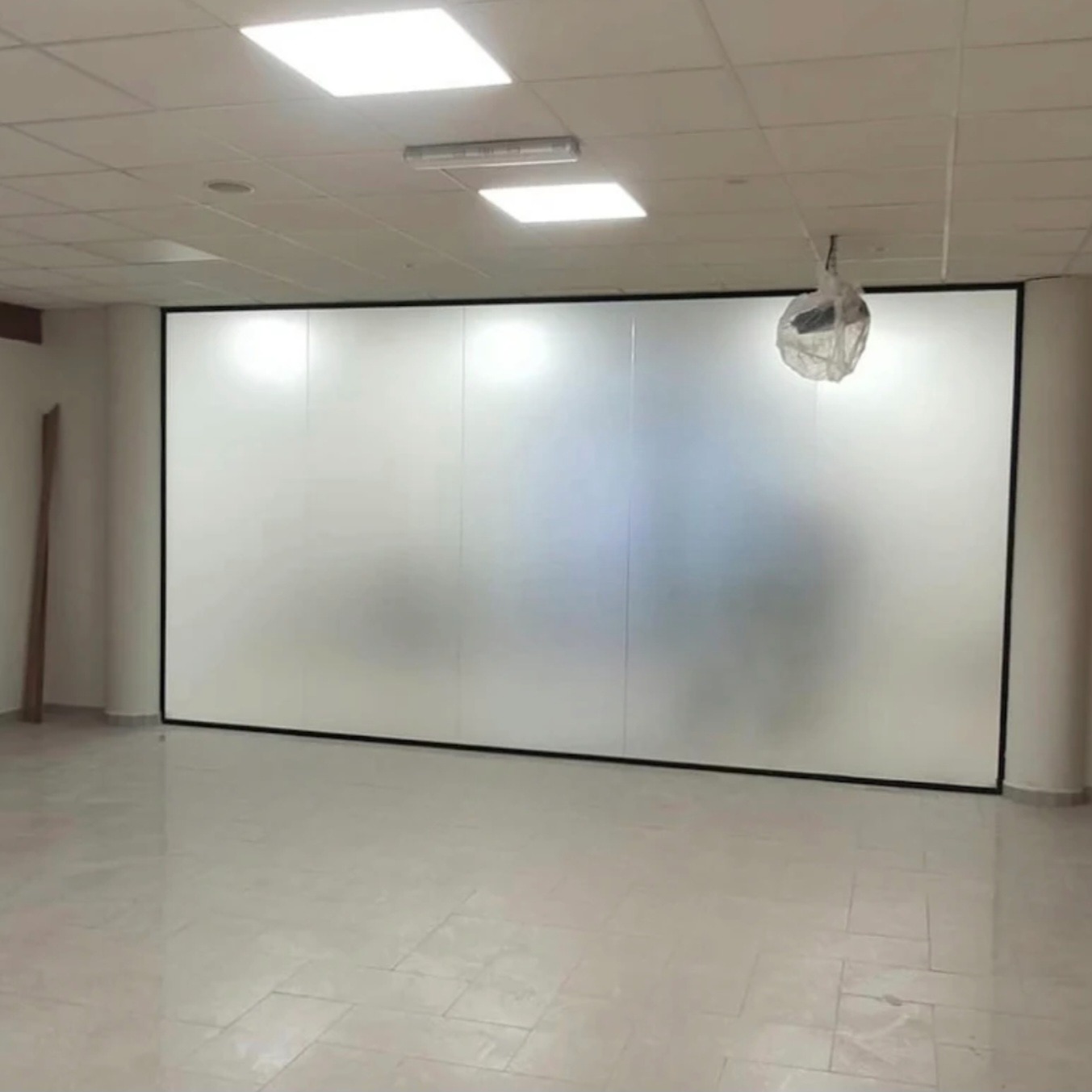 Duże, matowe panele szklane z czarną ramą, tworzące ścianę działową w jasnym pomieszczeniu z kafelkową podłogą. Widoczne elementy oświetlenia sufitowego.