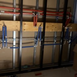 Filip Kliś Hydro-Bud - Instalacja wodna w trakcie montażu: czerwone i niebieskie rury PEX przymocowane do ściany z metalowymi profilami. Widoczne narzędzia i otwory w podłodze.