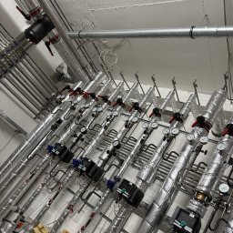 Filip Kliś Hydro-Bud - Rzędy izolowanych rur z zaworami i manometrami w kotłowni, widok z dołu. Elementy instalacji grzewczej w budynku, srebrna izolacja termiczna.