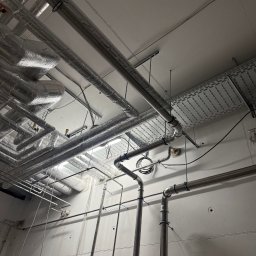 Filip Kliś Hydro-Bud - Srebrne rury instalacji przemysłowej pod sufitem, niektóre z izolacją, prowadzone wzdłuż ściany z widocznymi mocowaniami i elementami oświetlenia.