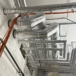 Filip Kliś Hydro-Bud - Srebrne, zaizolowane rury instalacji grzewczej pod sufitem z cegły, z widocznymi przewodami i elementami mocującymi. Układ rur tworzy skomplikowaną sieć.