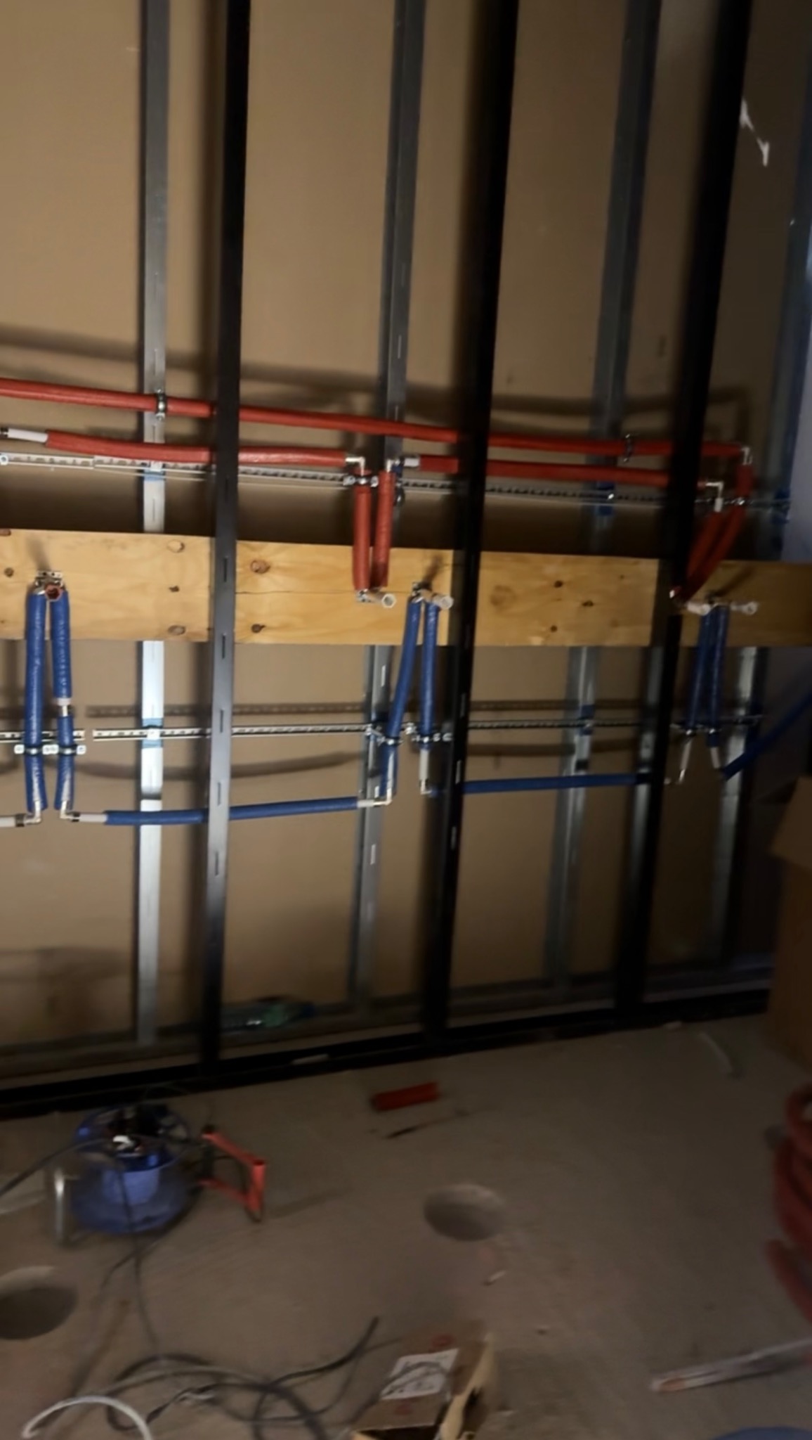 Instalacja wodna w trakcie montażu: czerwone i niebieskie rury PEX przymocowane do ściany z metalowymi profilami. Widoczne narzędzia i otwory w podłodze.