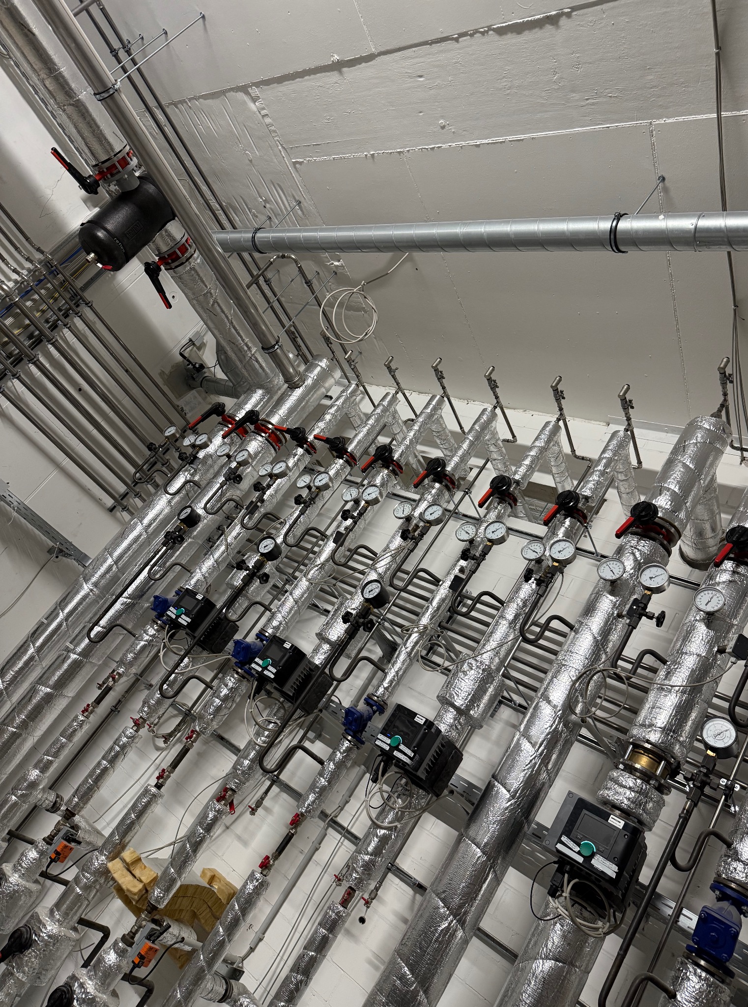 Rzędy izolowanych rur z zaworami i manometrami w kotłowni, widok z dołu. Elementy instalacji grzewczej w budynku, srebrna izolacja termiczna.