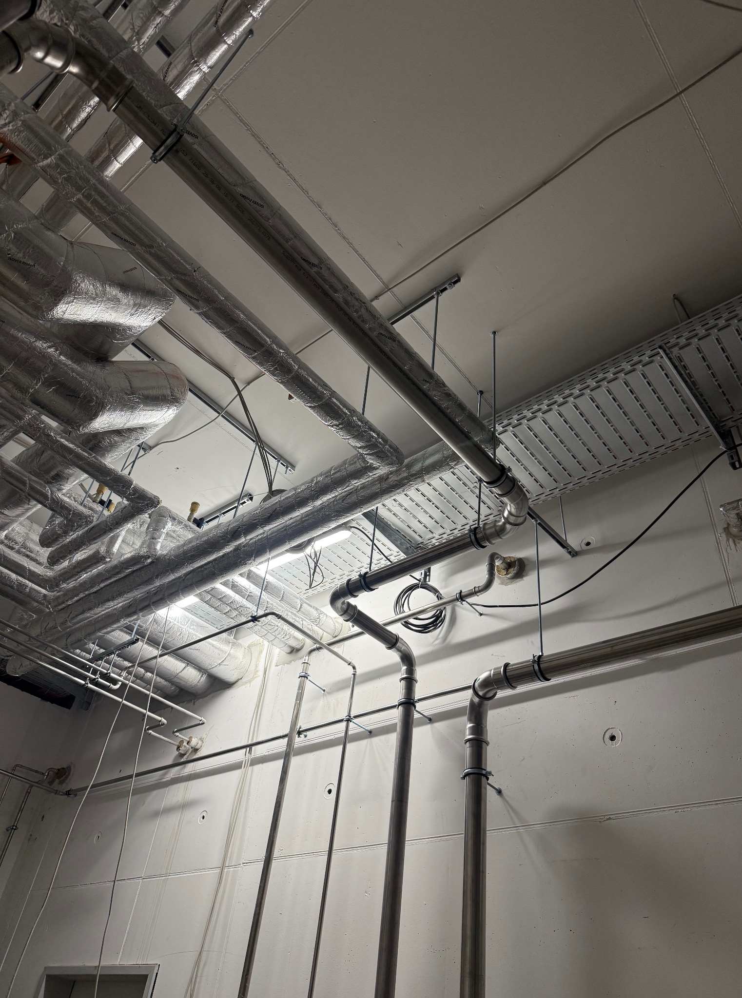 Srebrne rury instalacji przemysłowej pod sufitem, niektóre z izolacją, prowadzone wzdłuż ściany z widocznymi mocowaniami i elementami oświetlenia.