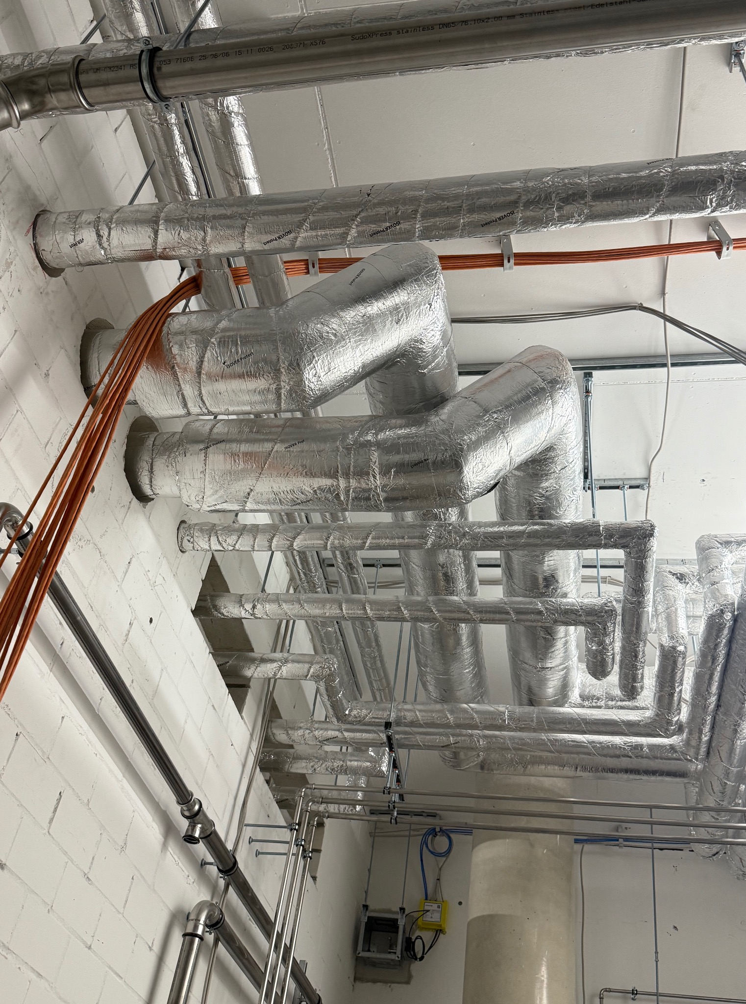 Srebrne, zaizolowane rury instalacji grzewczej pod sufitem z cegły, z widocznymi przewodami i elementami mocującymi. Układ rur tworzy skomplikowaną sieć.