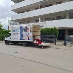 IMIXI Przeprowadzki Kielce - Biała furgonetka firmy IMIXI z otwartą rampą załadunkową, na której leżą czerwone tekstylia, zaparkowana przed nowoczesnym apartamentowcem, gotowa do przewozu.