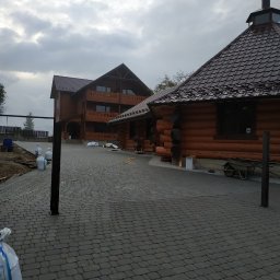 BUDZELONYRAJ - Ułożona kostka brukowa na podjeździe do drewnianego domu z bali, widoczne worki z materiałem i taczka. Pochmurne niebo w tle.