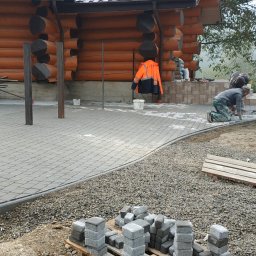 BUDZELONYRAJ - Układanie szarej kostki brukowej na posesji z drewnianym domem. Widoczni pracownicy w trakcie pracy, stosy kostki na palecie i pomarańczowa kurtka robocza.