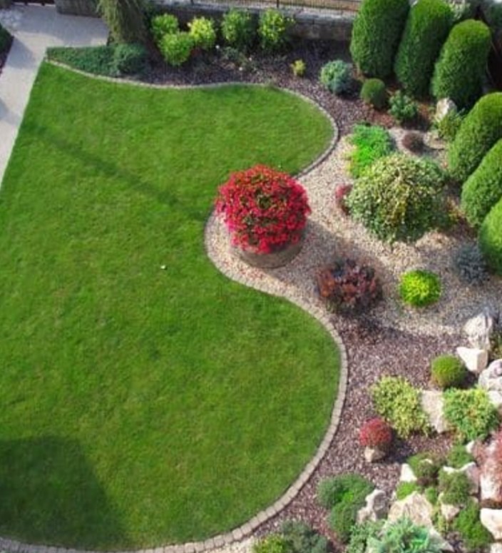 Zadbany ogród z równo przystrzyżonym trawnikiem, ozdobnymi krzewami i ścieżką wysypaną kamieniami. Centralnie umieszczona donica z czerwonymi kwiatami.