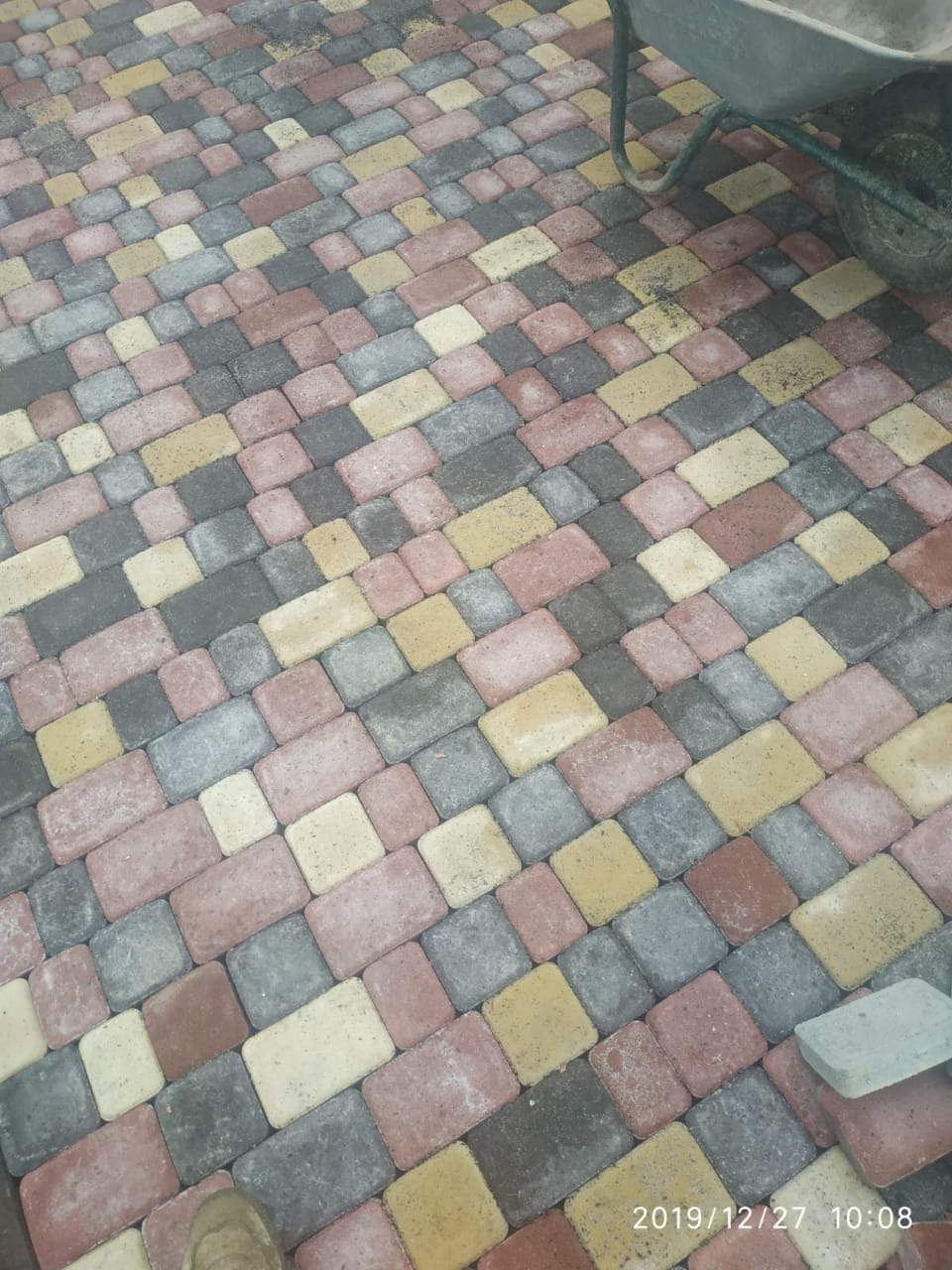 Kolorowa kostka brukowa ułożona w jodełkę, widoczna taczka w prawym górnym rogu i fragment buta w lewym dolnym rogu. Różne odcienie szarości, czerwieni i żółci.