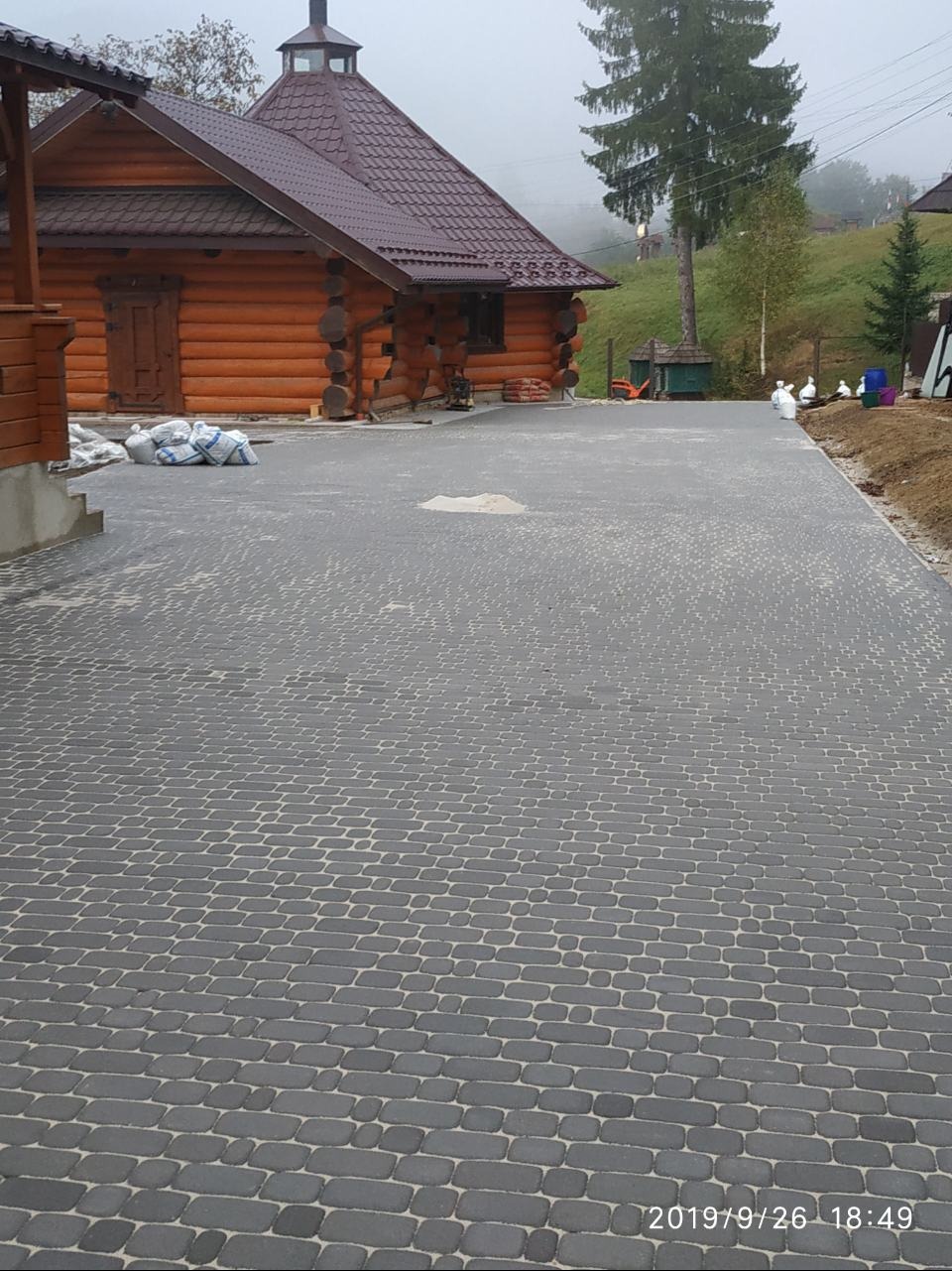 Ułożona szara kostka brukowa przed drewnianym domem z brązowym dachem. W tle zieleń i mgła. Widoczne worki z materiałem budowlanym.