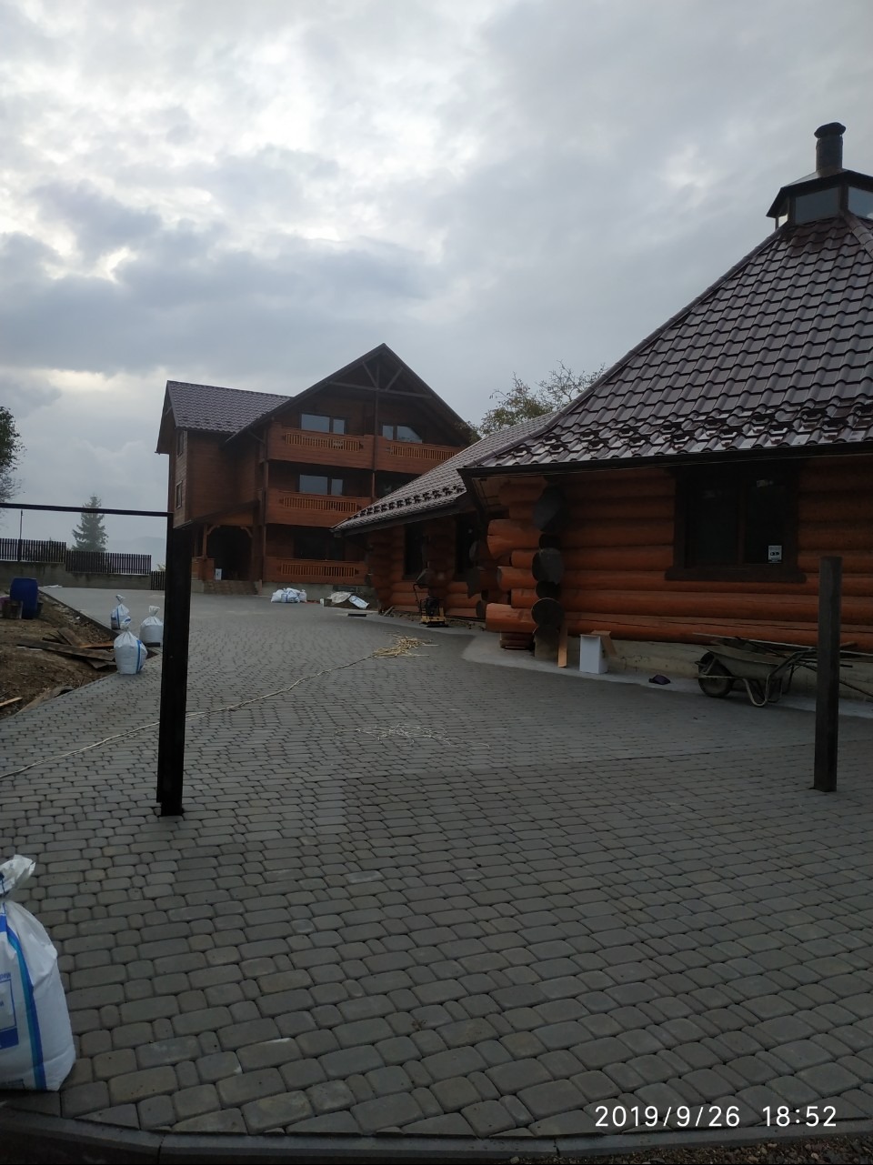 Ułożona kostka brukowa na podjeździe do drewnianego domu z bali, widoczne worki z materiałem i taczka. Pochmurne niebo w tle.