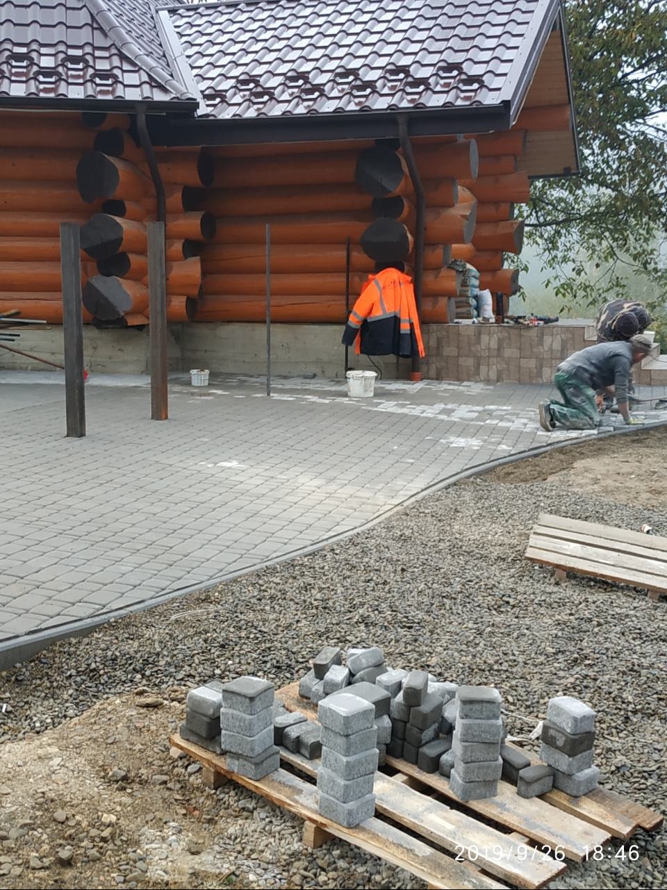 Układanie szarej kostki brukowej na posesji z drewnianym domem. Widoczni pracownicy w trakcie pracy, stosy kostki na palecie i pomarańczowa kurtka robocza.