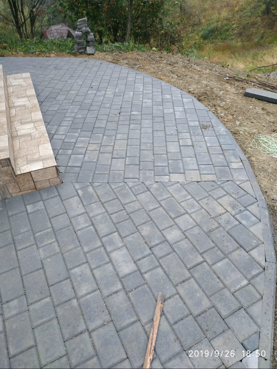 Ułożona szara kostka brukowa na podwórku, widok z góry. Fragment schodów z boku. W tle ziemia i roślinność. Data wykonania zdjęcia w prawym dolnym rogu.