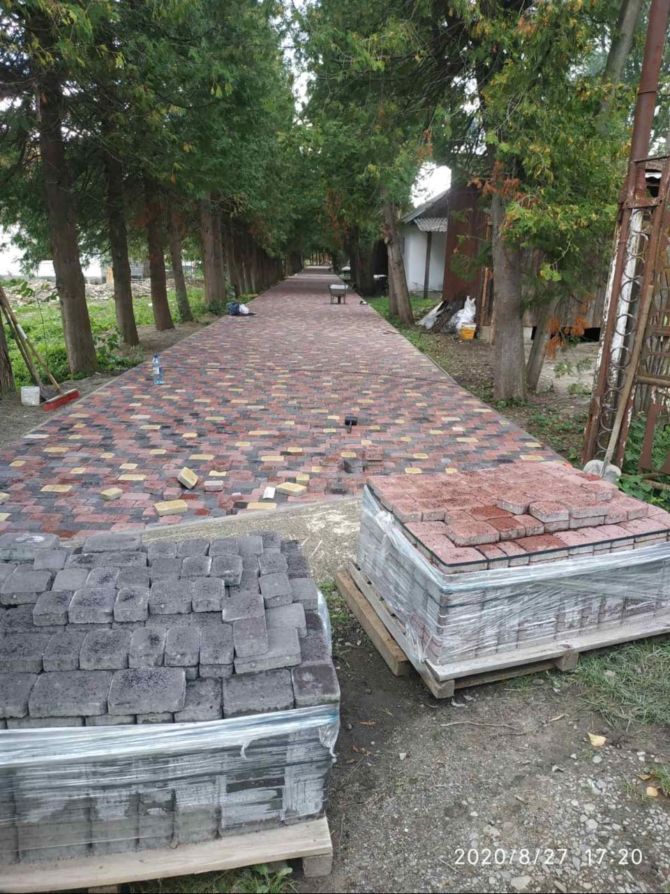 Aleja w trakcie układania z kostki brukowej w odcieniach czerwieni, żółci i szarości. Ułożona częściowo, z paletami kostki na pierwszym planie i drzewami wzdłuż ścieżki.