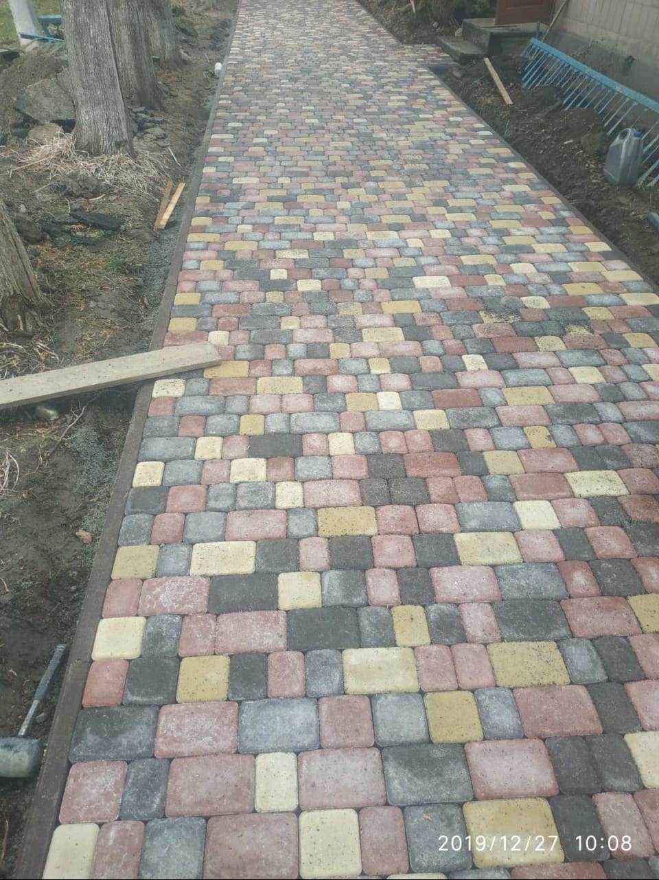 Kolorowa ścieżka z kostki brukowej prowadząca wzdłuż drzew, z widocznymi narzędziami i datą wykonania. Różnorodność barw kostki i perspektywa nadają unikalny charakter.