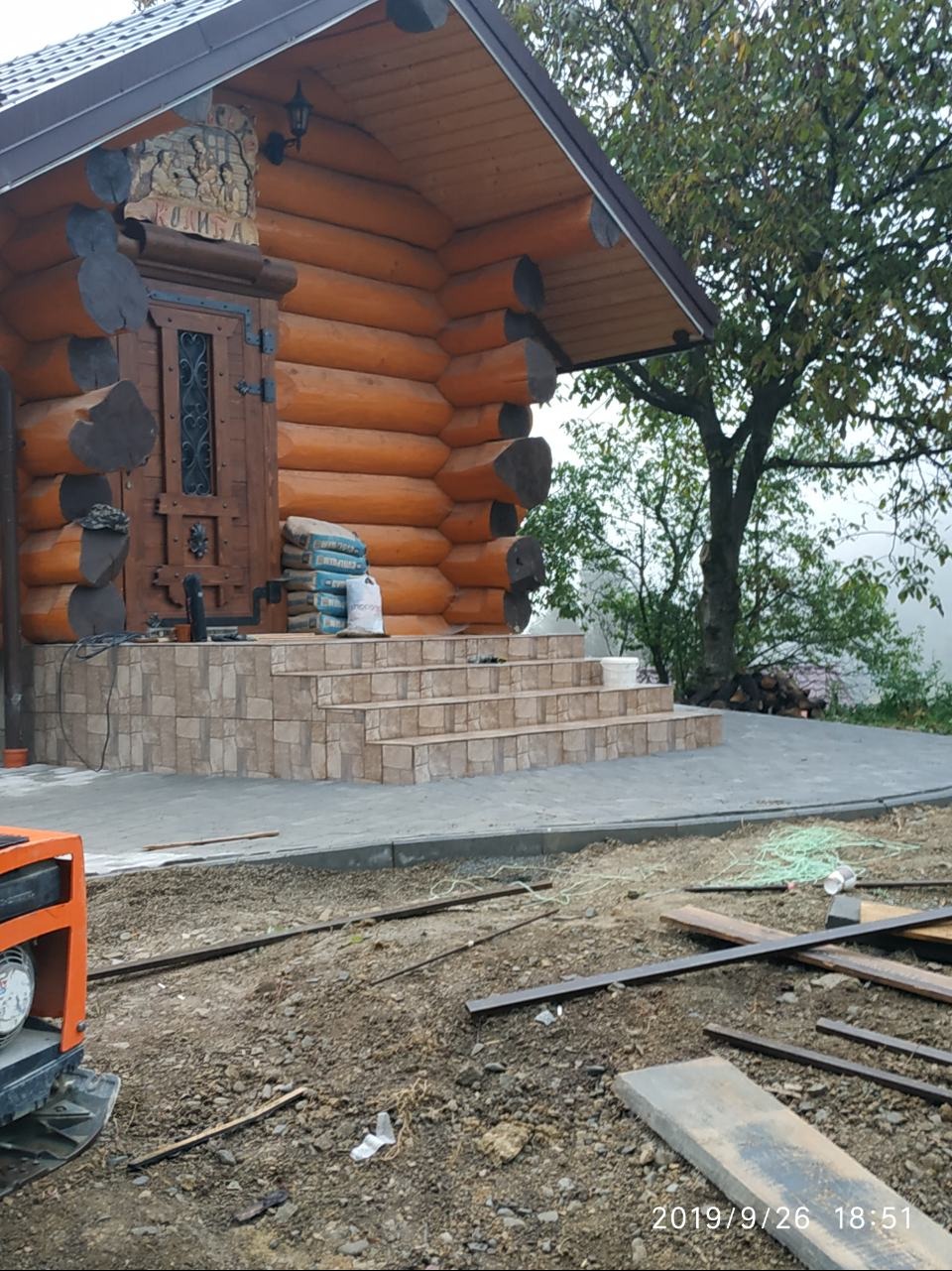 Wejście do drewnianego domu z kostki brukowej, widoczne worki z cementem i teren w trakcie prac wykończeniowych. Ujęcie z dołu, częściowo widoczny generator.