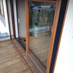 Glatboard Wojciech Glatman - Drewniane okno przesuwne z czarnym akcentem na balkonie z desek. Widoczne detale konstrukcyjne i odbicie otoczenia w szybie.