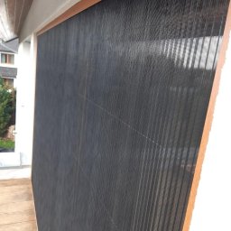 Glatboard Wojciech Glatman - Zamknięta, czarna moskitiera ramkowa z drewnianym obramowaniem, zamontowana na balkonie. Widoczne elementy konstrukcyjne i siatka. Ujęcie z bliska, z boku.