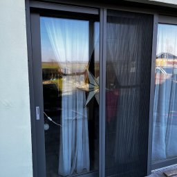 Glatboard Wojciech Glatman - Szara, przesuwna moskitiera plisowana na oknie tarasowym, widok z zewnątrz. W tle ogród i dom sąsiada, w oknie odbicie światła i dekoracja w kształcie gwiazdy.