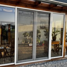 Glatboard Wojciech Glatman - Zasłonięte, przesuwne drzwi tarasowe z siatką przeciw owadom w jasnej ramie. Widoczne rośliny doniczkowe i meble wewnątrz przeszklonej werandy.