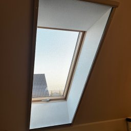 Glatboard Wojciech Glatman - Zamknięta moskitiera w oknie dachowym, widok na dach budynku i niebo. Delikatna siatka moskitiery zapewnia ochronę przed owadami.