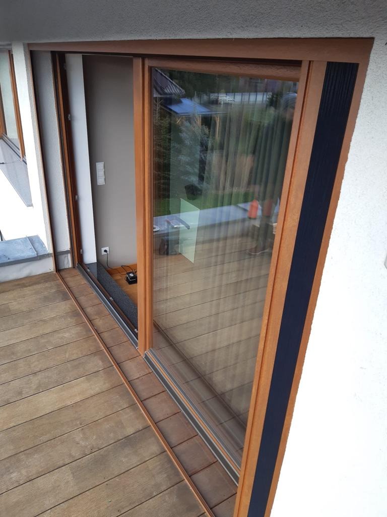 Drewniane okno przesuwne z czarnym akcentem na balkonie z desek. Widoczne detale konstrukcyjne i odbicie otoczenia w szybie.