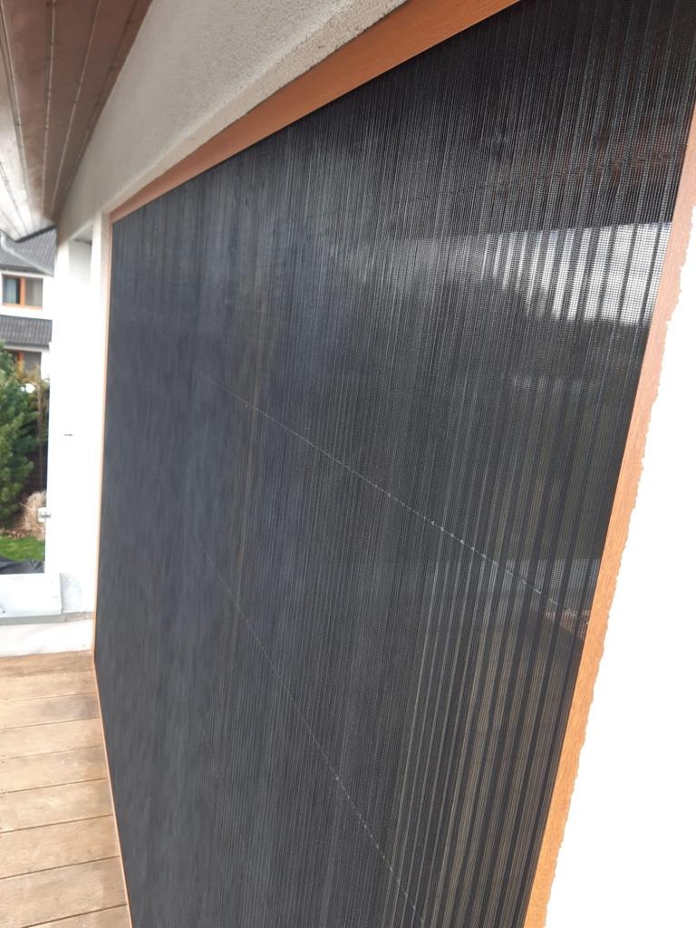 Zamknięta, czarna moskitiera ramkowa z drewnianym obramowaniem, zamontowana na balkonie. Widoczne elementy konstrukcyjne i siatka. Ujęcie z bliska, z boku.