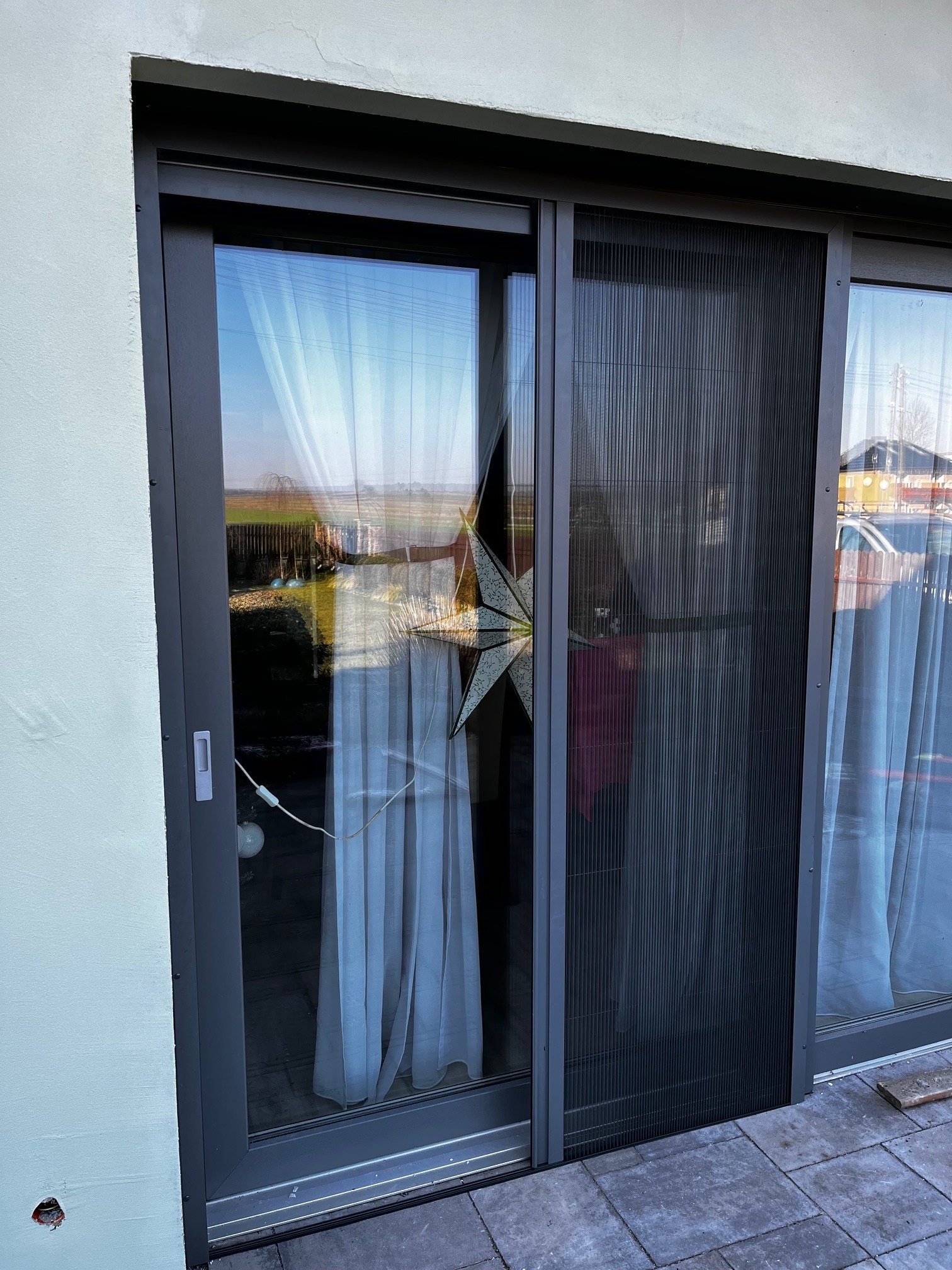 Szara, przesuwna moskitiera plisowana na oknie tarasowym, widok z zewnątrz. W tle ogród i dom sąsiada, w oknie odbicie światła i dekoracja w kształcie gwiazdy.