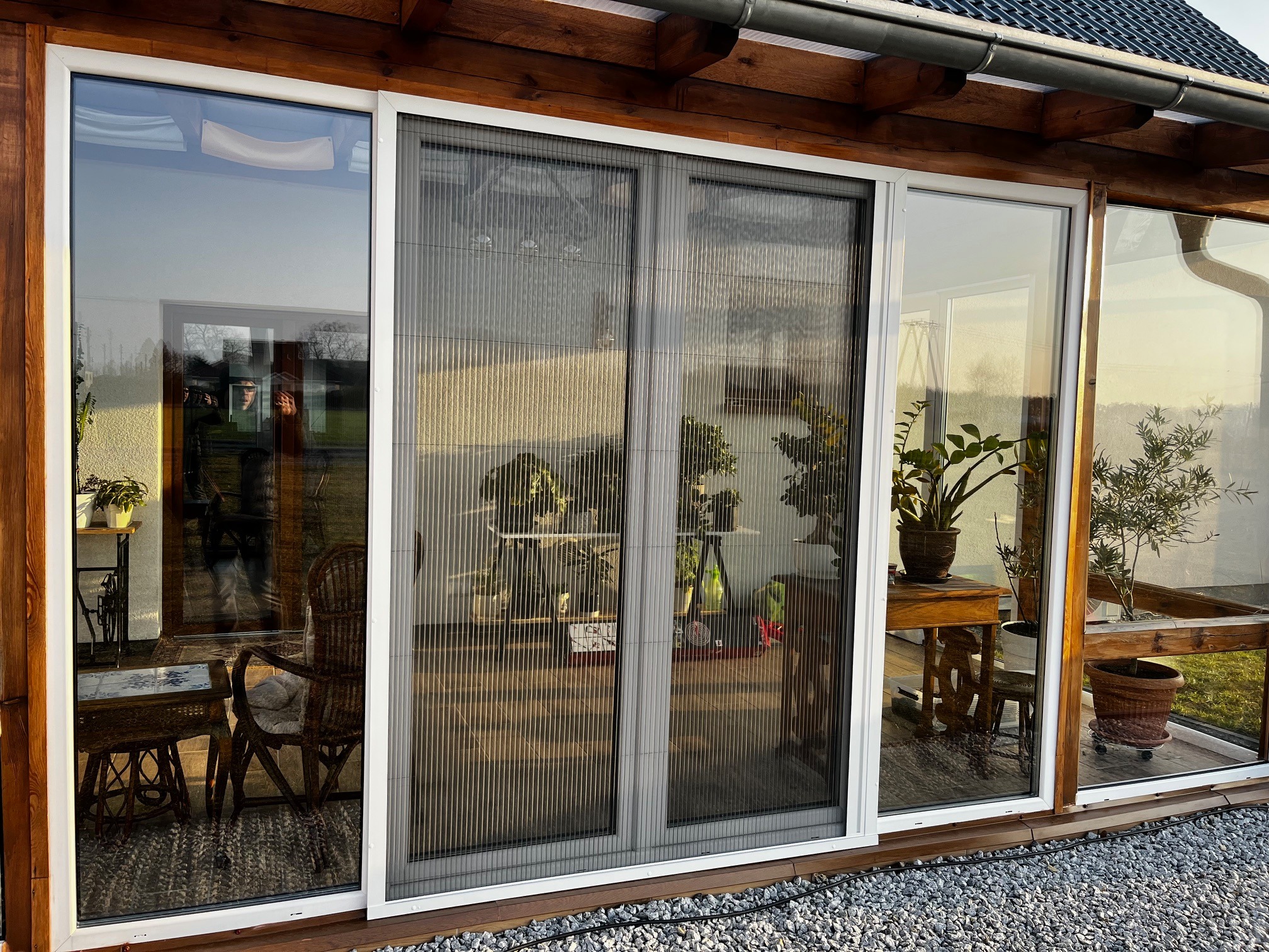 Zasłonięte, przesuwne drzwi tarasowe z siatką przeciw owadom w jasnej ramie. Widoczne rośliny doniczkowe i meble wewnątrz przeszklonej werandy.