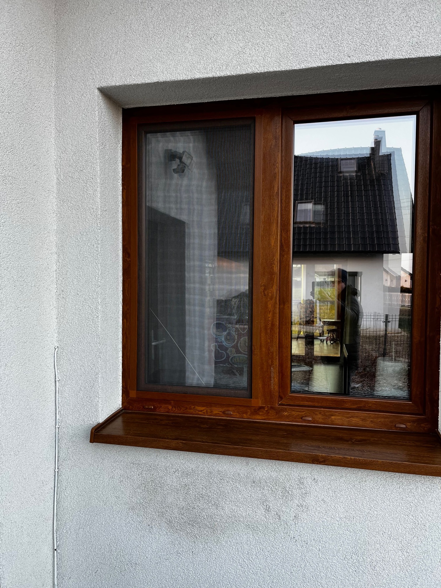 Okna z brązową ramą i moskitierą w białym budynku. Widoczne odbicie otoczenia w szybie okiennej. Detal moskitiery w oknie.