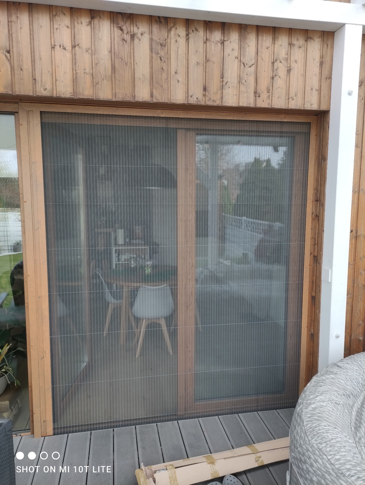 Złożona moskitiera plisowana w drewnianej ramie, zamontowana w drzwiach tarasowych. Widoczny fragment tarasu i wnętrze domu z meblami.