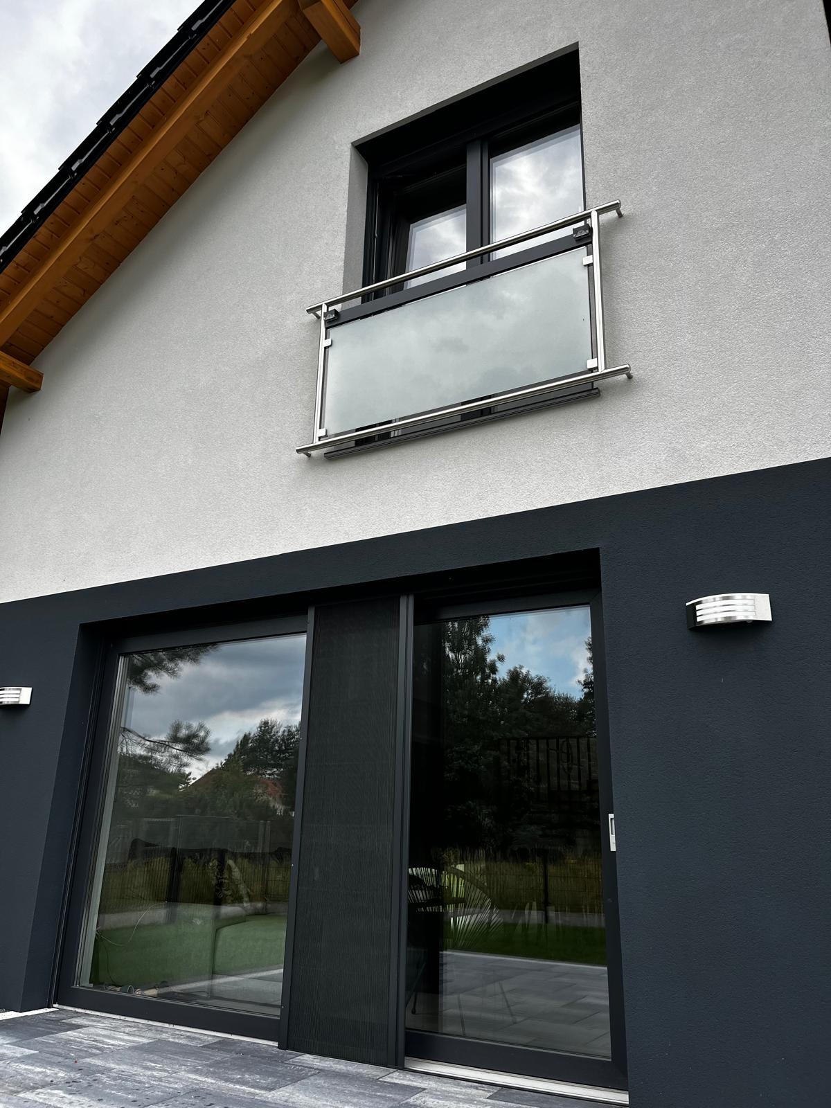 Nowoczesny dom z dużymi oknami i szarą elewacją. Na oknach zamontowane są moskitiery w kolorze ramy okiennej. Widoczny balkon z szklaną balustradą.