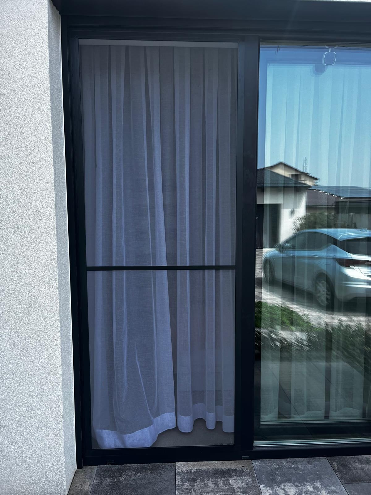 Zamknięte okno z moskitierą w czarnej ramie, przez które widać firankę i odbicie samochodu na zewnątrz. Widok z bliska na elewację budynku.