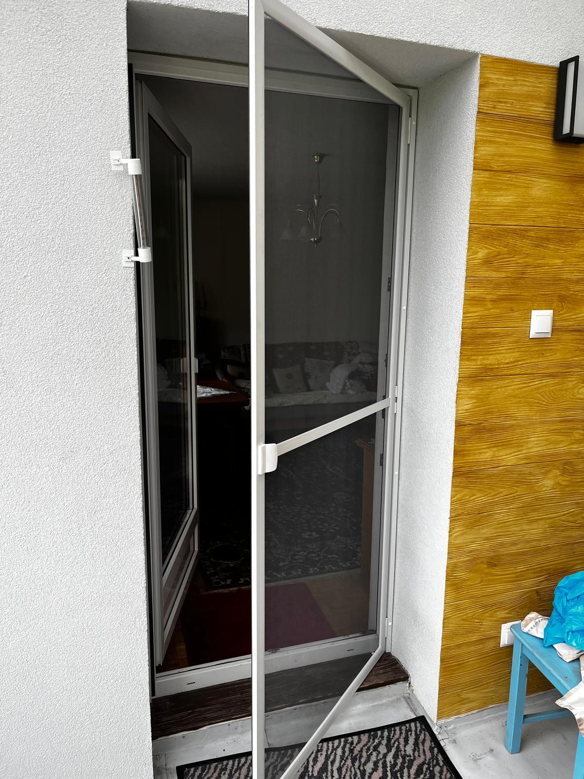 Otwarta moskitiera ramkowa w drzwiach balkonowych, widok z zewnątrz. Biała rama, siatka w kolorze grafitowym. Na zewnątrz widoczna drewniana boazeria i fragment niebieskiego krzesła.