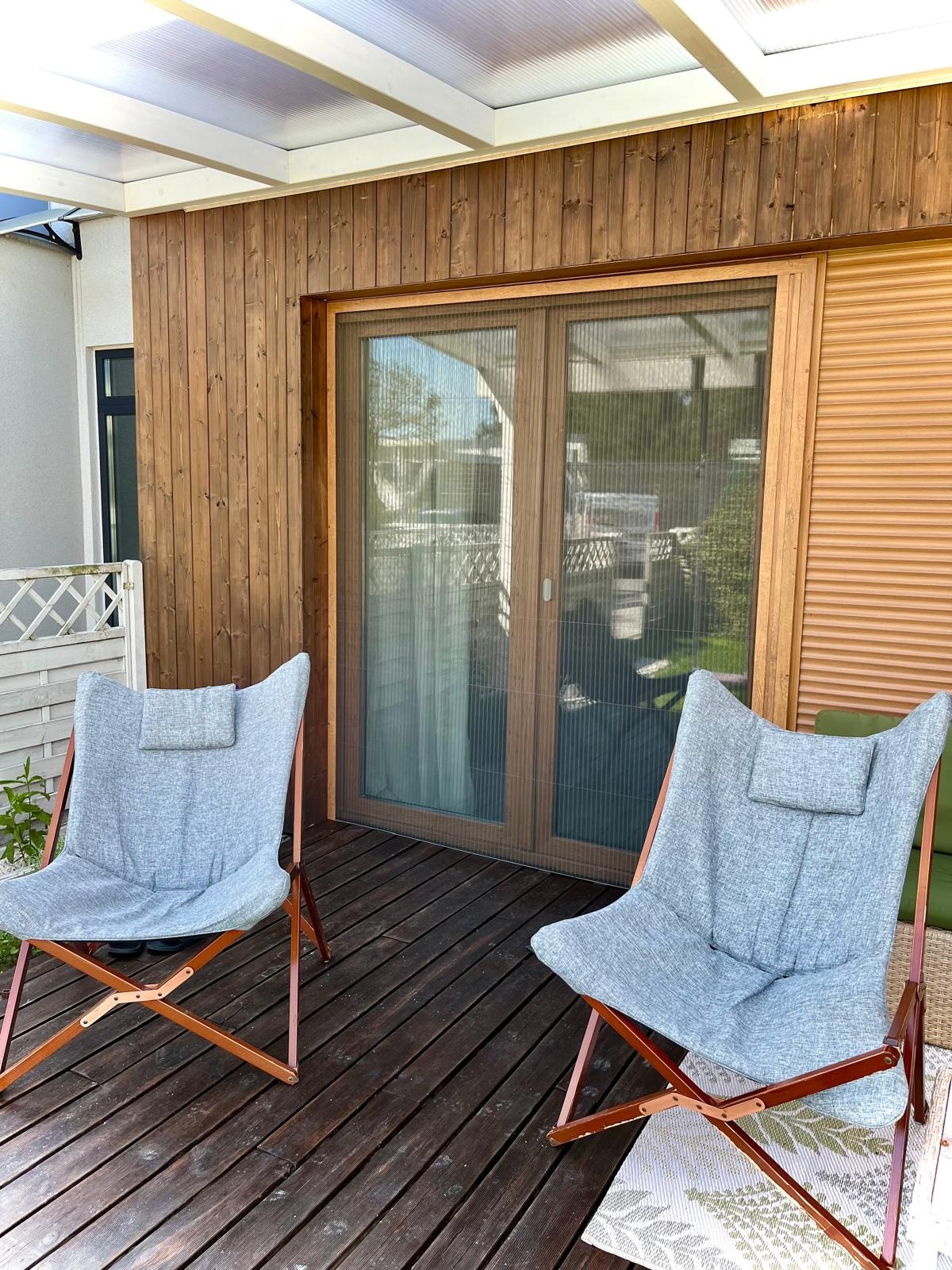 Zewnętrzna moskitiera przesuwna na dwuskrzydłowych drzwiach tarasowych, widok z zewnątrz, z dwoma krzesłami na pierwszym planie i drewnianą elewacją budynku.